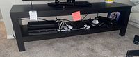 Front view of the black bi-level TV stand showing top surface and lower shelf with various electronics (not included in sale)