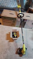 Full view of Ryobi weed eater with shaft and blade guard, with box on ground
