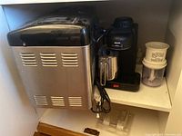 Black & Decker bread maker and Krups cappuccino maker side by side on a shelf with a small white accessory next to the cappuccino maker