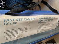 Blue carrying case for Caravan Fast Set Canopy 10'x10' with text detailing features including easy folding steel frame with telescopic legs, deck feet for stability, fabric made of 100% polyester, and powdered coated steel frame dimensions.