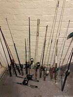 Photo shows a collection of more than a dozen fishing rods with attached reels lined up against a concrete wall. The rods vary in length and handle style.