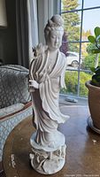 Full view of white porcelain goddess figurine standing on lotus base near window light.