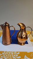 Front view showing two copper jugs side by side on table, one tall and slender with lid and handle, the other round with curved spout and handle