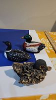 Three duck items displayed: a mallard duck decoy, a loon piggy bank, and a clay duck sculpture on a white and blue background with part of a yellow floral fabric and a ruler for scale.