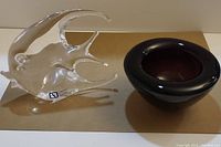 Clear Murano glass fish sculpture next to small dark purple glass bowl on gold surface