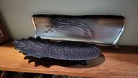 Two metal trays displayed on a wooden surface, showing a textured pineapple-shaped tray and a rectangular etched tray with thistle design