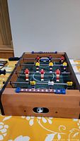 Wooden tabletop foosball game setup showing players in red, blue, yellow, and white uniforms on rods with black handles and green field with white markings.
