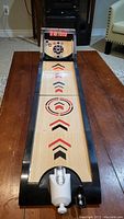 Front view of the full skeeball tabletop game showing the wooden play surface with target rings and arrows, framed by black plastic edges, on a brown table.