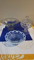 Three vintage glass candy dishes placed on a blue cloth: lidded hobnail dish, blue dish with openwork rim, and large footed dish.