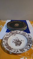 Stack of three decorative ceramic wall plates showing the front of one with brown floral urn pattern and another with dark green and gold pattern.