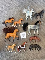 Top-down view of 10 toy horse figures of various colors and poses laid out on carpeted floor. Includes two characters inspired by TV show Spirit, one white with flowing mane/tail and one dark brown.