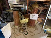 Two antique brass carafes displayed on a table, showing one with a broken lid hinge and the other tall carafe with a spout and handle.