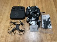 Top view of Sharper Image DX-2 drone, Zenfolt x-Pack 7 drone, their controllers, spare propellers, and manuals on a wood floor.