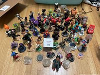 Photo showing a large variety of Disney Infinity character figures arranged on a wooden floor, including characters from Marvel, Pixar, Star Wars, and Disney franchises. Some figures have parts missing or bases are detached.