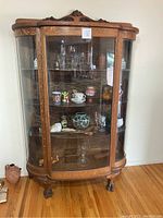 Full view of the antique wooden china cabinet with curved glass front and side panels, containing glassware and other small items for scale.