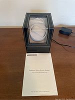 Black watch winder box with clear acrylic top and sides, showing cushioned beige watch holder inside. Power cord connected and instruction manual placed in front.