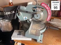 Photo showing Delta 10-inch power miter saw with blade guard and dust collection bag, placed on a wooden table with the user's inventory tag visible.
