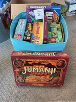 Photo showing a blue basket filled with multiple boxed board games with a Jumanji board game box in front on the carpeted floor.