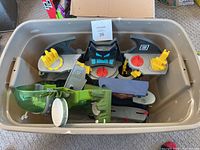 Plastic bin containing disassembled Batman and Superman Imaginext playset parts. Features grey, black, yellow, and red colored pieces including Bat-symbol parts and wing shaped pieces.