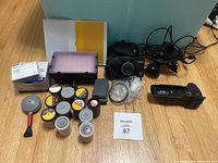 Photo showing collection of 35mm film rolls, camera filters, Canon Prima Zoom 70F camera with case, LED light panel, CANON Vertical Grip VG10, DV batteries, dashcam and cleaning brush on wooden surface