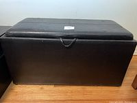 Front and side view of black faux leather storage bench showing rectangular shape and hinged lid with pull handle.