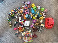 Full view of various toys and figurines laid out on carpet, showing the diversity of characters including Olivia's red car, Smurfs, and new Lalaloopsy doll in original packaging.