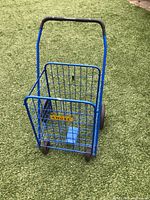 Front view of blue Angel brand collapsible cart showing the wireframe with brand label, black handle grip, and front small wheels on artificial grass.