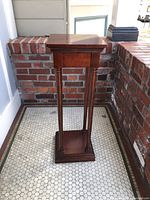 Full view of the wooden pedestal stand placed against a brick wall showing the overall structure and height.