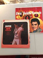 Two Elvis Presley LP albums placed on a white surface; one cover showing Elvis performing in white jumpsuit, the other bright red with gold record images and Elvis' portrait.