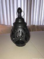 Front view of dark grey metal candleholder with ornate cutout design and hanging ring on top.