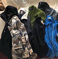 Full view of seven jackets on cloth background revealing overall variety in color and fabric including camouflage, black, green, blue and leather textures