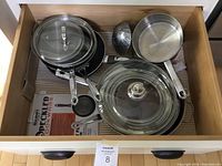 Drawer with multiple frying pans, saucepans, glass lids, double boiler parts, and steamer basket arranged inside.