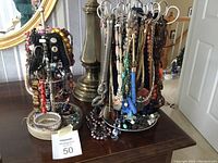 Two black metal jewelry stands holding multiple colorful costume necklaces and bracelets, displayed on a wooden table near a mirror and lamp.