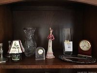 Full lot showing candlesticks, small lamp with floral shade, two glass vases, Waterford clock, female figurine, Howard Miller clock, and silver-tone tray in wooden cabinet
