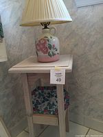 Overall view of light natural colored wooden square decorative table with jug-shaped lamp on top and ceramic plate on lower shelf.