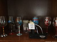 Shelf with multiple painted themed wine glasses with black and white checkered, red, and candy cane striped stems, each decorated with Broadway or Italy motifs.