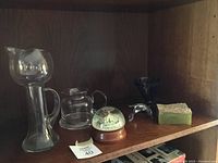 Photo showing six decorative items on a wooden shelf including two glass pitchers, snow globe, glass dolphin, tall glass vase, and stone container with lid.