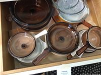 Top-down view showing collection of brown glass stove top pans with lids, white handled glass pan, and clear glass Corning Ware sauté dishes.