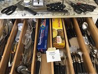 View inside wooden kitchen utensil drawer showing organized black handled cutlery including knives, forks, spoons, and salad forks, plus assorted kitchen tools and wrap boxes.