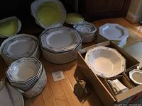 Full set of Nikko Classic Collection dinnerware arranged on floor and in boxes. Visible plates, bowls, and serving pieces with blue trim and plain varieties.