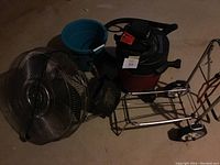 Wide shot of entire lot showing shop vac, oscillating fan, metal cart, blue bucket, and dustpan on dark floor.