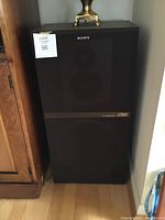 Full front view of a single brown floor-standing Sony speaker with black grill and branding, showing size relative to nearby furniture.