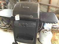 Front view of the Thermos gas grill showing the grill lid, side tables, front control panel, and attached propane tank beside it.