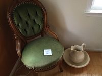 Full view of Victorian green chair next to off-white pitcher and basin on wood floor.