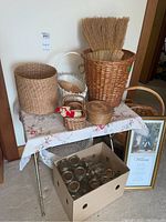Table with multiple woven baskets in various sizes and styles, two straw whisk brooms inside largest basket, small straw figurine, framed print leaning against table, and box with clear glass canning jars.