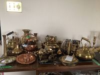 Full view of mixed brass and copper items displayed on wooden surface including teapots, vases, kettles, candle sconces, and plates.