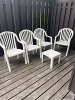 Four white stackable outdoor plastic deck chairs with vertical slats on back and armrests, arranged on a wooden deck next to a black fence, along with a small white plastic table with a slatted top.