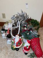 Photo shows various ceramic holiday figures including two Santa candle holders, snowmen, and elf, alongside baskets, pine cones, gold star, wreaths, and box of candles arranged together.