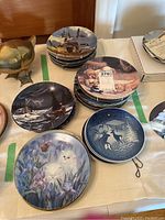 Photo showing multiple stacks of decorative and collectible plates with various wildlife and animal scenes including puppies, cats, and birds.