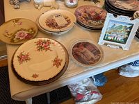 Table display of assorted floral and themed decorative plates with floral and seasonal patterns and floral arrangements.